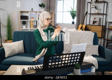 Junge muslimische Frau in Hijab spielt Flöte auf hellen Wohnzimmer zu Hause. Mädchen sitzt auf dem Sofa mit einem Musikinstrument drinnen. Hübsche junge arabische Frau studiert Flöte spielen. Stockfoto