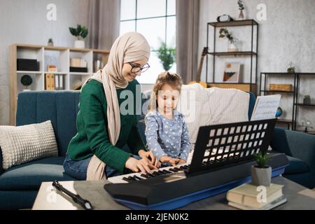 Kleines nettes Mädchen mit Musik muslim Lehrer mit Unterricht am Klavier an der Musikschule. Lächelnde junge arabische Frau, die auf ein kleines Mädchen schaut, das auf dem Sofa sitzt und im Klassenzimmer zusammen Klavier spielt. Stockfoto