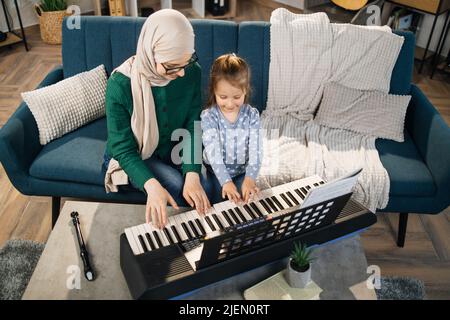 Porträt einer muslimischen Frau in Hijab und ihrer niedlichen kleinen Tochter, die zu Hause Klavier spielen lernt. Lehrerin, die hübsches Mädchen unterrichtet, um im Klassenzimmer Klavier zu spielen. Stockfoto