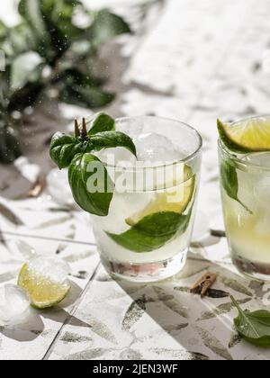 Zwei Sommercocktails mit Limette und Basilikum in Gläsern schließen auf weiß bedruckten Fliesen. Vertikale Ausrichtung Stockfoto