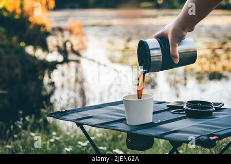 Gießen Sie heißen schwarzen Kaffee in wiederverwendbaren Bambusbecher auf Campingtisch im Freien während der frühen Morgenstunden. Frisch gebrühten Kaffee in der Natur zubereiten Stockfoto