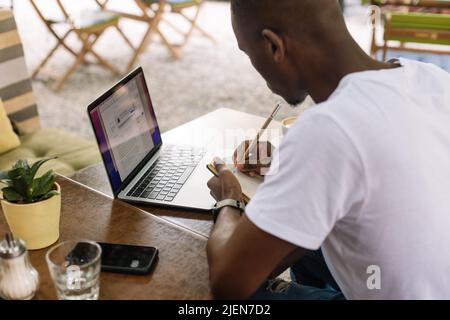 Ein aus mehreren Rassen zugeschnittenes Geschäftsmann, der mit einem Laptop unterwegs war, Notizen im Arbeitsbuch machte, Kaffee im Straßencafé trank. Hoher Winkel Stockfoto