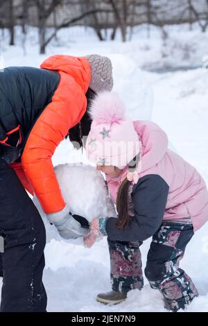 Kleine Tochter und Mutter mittleren Alters machen einen mittleren Teil des Schneemanns zusammen auf dem Hinterhof am Abend mit Bäumen im Hintergrund. Eltern verbringen Zeit Stockfoto