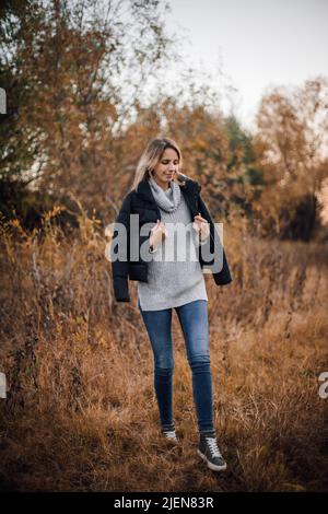 Porträt einer jungen Frau, die auf einer Wiese im Wald auf Gras läuft und lächelnd mit einer schwarzen Jacke auf den Schultern und bedeckten Bäumen nach unten blickt Stockfoto