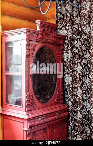 Roter hölzerner Vintage-Schrank mit geschnitzten Ornamenten steht in der Ecke des ländlichen Holzhauses zwischen Fenstern in der Nähe von Vorhang. Stockfoto