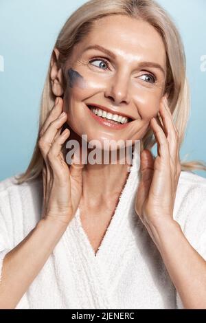 Vertikale lächelnde ältere Frau, die ihre gesunde Haut mit Gesichtscreme, Gesichtsmaske und Peeling auf der Wange behandelt, die auf blauem Hintergrund steht Stockfoto