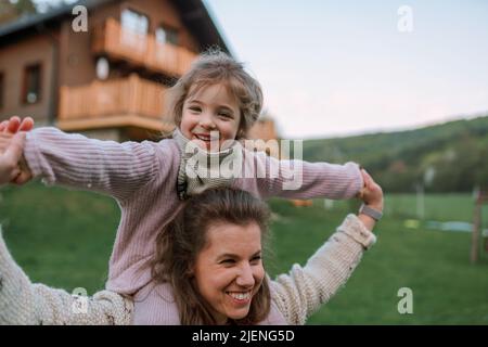 Glückliche Mutter mit kleiner Tochter auf Huckepack laufen und Spaß zusammen im Garten in der Nähe ihres Hauses. Stockfoto