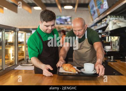 Glücklicher Kellner mit Down-Syndrom, der Kaffee mit Hilfe seines Kollegen im Café serviert. Stockfoto