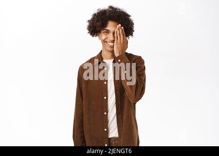 Porträt eines lächelnden hispanischen jungen Mannes mit afrohaarigen Haaren, bedeckt die Hälfte des Gesichts, Augengeste, stehend über weißem Hintergrund Stockfoto