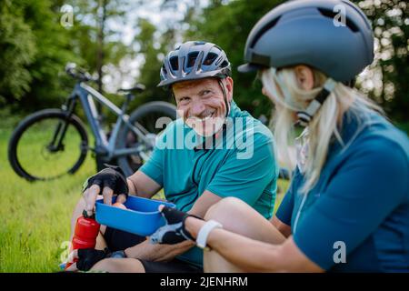 Aktives Seniorenpaar, das sich nach einer Fahrradtour im Sommerpark ausruhte, auf Gras saß und einen Snack zu sich nahm. Stockfoto