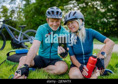 Aktives Seniorenpaar, das sich nach einer Fahrradtour im Sommerpark auf dem Rasen und mit dem Smartphone ausruhte. Stockfoto