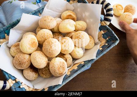 Käsebrot in einem rechteckigen Korb und ein Teil der Hand ist unscharf und nimmt ein Brötchen auf einem Teller in der rechten Ecke auf Stockfoto