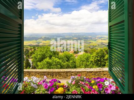 Blick durch ein offenes Fenster mit Fensterläden auf die toskanische Landschaft und die mittelalterliche Altstadt von San Gimignano, Italien. Stockfoto