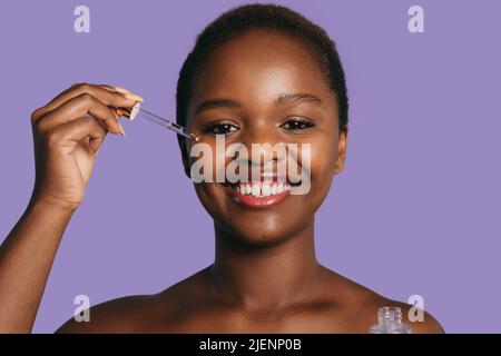 Lächelnde Afro-Frau, die feuchtigkeitsspendende Gesichtsserum auf ihre Haut aufgetragen, isoliert auf violettem Hintergrund. Schöne schwarze Frau. Natürliche Schönheit. Gesichtsbehandlung Stockfoto