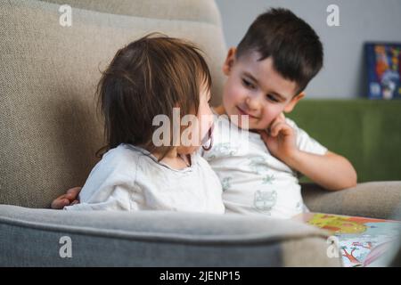 Portrait von zwei kleinen Kindern, die einen Roman lesen, lächeln und einander zuschauen, während sie auf dem Sofa sitzen. Kaukasische Kinder ruhen sich aus und genießen die Freizeit Stockfoto