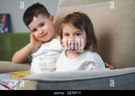 Bruder und Schwester sitzen auf dem Sofa und posieren beim Lesen des Buches zu Hause. Lustige Märchengeschichte. Gemeinsame Freizeit am Wochenende. Stockfoto