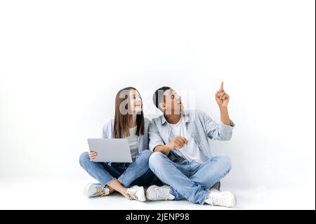 Die hübsche kaukasische Frau und der gutaussehende hispanische Mann, ein positives Paar, sitzen mit einem Laptop auf dem Boden auf weißem, isoliertem Hintergrund und schauen zur Seite, der Typ zeigt mit der Hand auf etwas Stockfoto