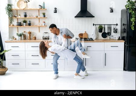 Ein junges, modernes, glückliches Paar verliebt, frisch verheiratetes Paar, stilvoll gekleidet, tanzen zu ihrer Lieblingsmusik zu Hause in der Küche mit stilvollem Interieur, lächeln und genießen es, Zeit miteinander zu verbringen Stockfoto