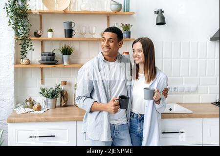 Glückliches multirassisches romantisches Paar, Brautpaar, stilvoll gekleidet, zu Hause in der Küche umarmt, Tassen Kaffee oder Tee in den Händen halten, wegschauen, träumen, lächeln, Morgenstunden miteinander verbringen Stockfoto