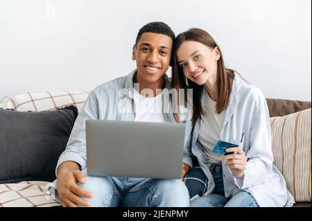 Online-Shopping, Technologie. Positive multirassische Paar verliebt, Ehepartner, sitzen auf dem Sofa in einem Wohnzimmer, mit Laptop und Kreditkarte für Online-Shopping, Lieferung, Blick auf die Kamera, lächelnd Stockfoto