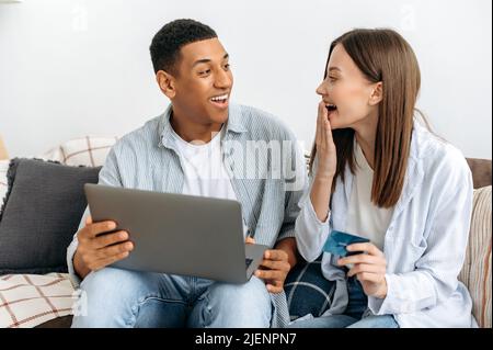 Aufgeregt glücklich stilvolle multirassische Paar verliebt, Ehegatten, sitzen auf dem Sofa in einem Wohnzimmer, mit Laptop und Kreditkarte für Online-Shopping, Lieferung, Blick erstaunt einander, lächelnd Stockfoto