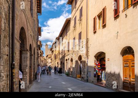 Touristen wandern durch die enge Hauptfußgängerzone voller Geschäfte und Cafés durch die mittelalterliche Hügelstadt San Gimignano, Italien, in der Toskana. Stockfoto