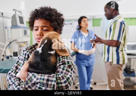 Junger afroamerikanischer Junge umarmt seinen Welpen und tröstet ihn nach einer schmerzhaften Impfung, sein Vater und sein Tierarzt diskutieren etwas vor dem Hintergrund Stockfoto