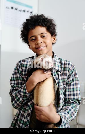 Vertikales mittleres Porträt eines fröhlichen afroamerikanischen Kindes, das sein liebliches Frettchen in der Kamera lächelt Stockfoto