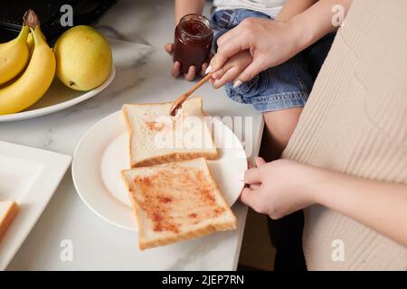 Die Hände der Mutter verteilen die Beerenmarmelade auf die Brotscheibe beim Kochen des Frühstücks Stockfoto