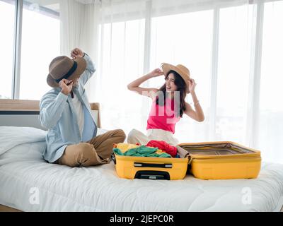 Paar Reisende, die sich auf die Reise vorbereiten. Junge asiatische Frau und Mann im Denim-Hemd tragen Strandhüte, während sie mit gelben Koffern auf dem Bett im Bett packen Stockfoto