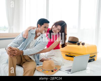 Die Freundin bettelt und überredet ihren Freund zu einem Sommerurlaub. Junge süße asiatische Frau und schöner Mann, der auf dem Laptop-Display auf Stockfoto