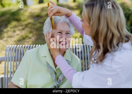 Pfleger hilft älteren Frauen, Haare zu kämmen und Frisur zu machen, wenn sie im Sommer auf der Bank im Park sitzen. Stockfoto