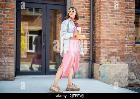 Stilvolle Frau geht mit Kaffeetasse in der Nähe von modernen Bürogebäude im Freien Stockfoto