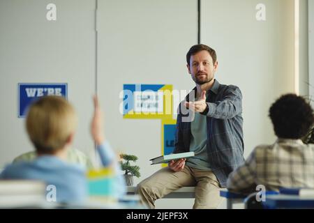 Porträt eines bärtigen männlichen Lehrers, der während des Unterrichts Fragen stellt, wobei die Kinder die Hände heben und den Raum kopieren Stockfoto
