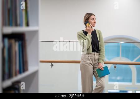 Frau mit Buch in den Händen, die in der modernen öffentlichen Bibliothek auf dem Mobiltelefon spricht Stockfoto