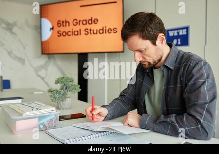 Porträt eines männlichen Lehrers, der im Klassenzimmer am Schreibtisch sitzt und die Hausaufgaben der Kinder bewertet Stockfoto