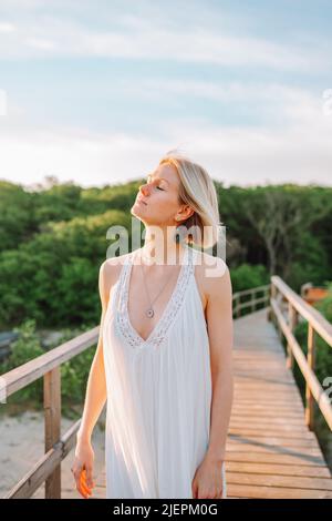 Porträt einer ruhigen Frau mittleren Alters mit hellen Haaren tragen lange weiße Licht Sommersundress, zu Fuß auf Holzbrücke im Wald an sonnigen Tag, luxuriös Stockfoto