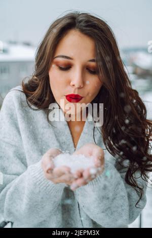 Porträt einer jungen attraktiven Frau mit langen, welligen, dunklen Haaren, Make-up in grauer Wolljacke, Schneeflocken aus den Handflächen, die nach unten schauen. Winter Stockfoto