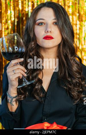 Porträt einer jungen ernsthaften Frau mit langen, welligen, dunklen Haaren, Make-up in schwarzem Hemd, Glas mit Rotwein auf goldenem Lametta-Hintergrund. Ein Großes Stockfoto