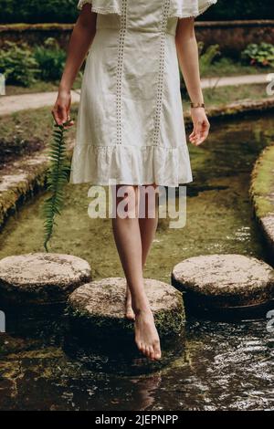 Frau mit Farn in den Händen, die auf Steinen steht und am Sommertag Wasser probiert. Sommerliches Naturkonzept Stockfoto