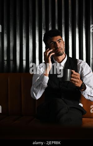 Ein hübscher und smarter asiatischer Geschäftsmann in formellem Outfit sitzt auf dem Ledersofa, nippt Kaffee und telefoniert mit seinem Geschäftspartner. Stockfoto