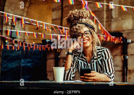 Porträt einer lächelnden Frau mit Afro-Frisur, Brille, Smartphone und Papiertasse, die in einer Coffeeshop-Aufnahme auf die Kamera schaut Stockfoto