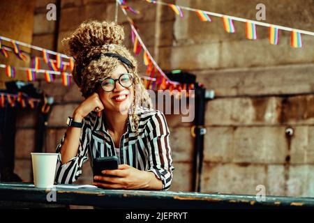 Porträt einer lächelnden Frau mit Afro-Frisur, Brille, Smartphone und Papiertasse, die in einer Coffeeshop-Aufnahme auf die Kamera schaut Stockfoto