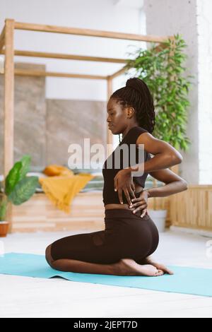 Vertikale multirassische Frau mit Dreadlocks Stretching, machen Yoga-Übungen, sitzen auf Beugen Beine. Rückenschmerzen nach dem Training Stockfoto