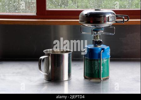 Ein kleiner Edelstahlkessel mit Klappgriffen heizt auf dem tragbaren Gasbrenner, der Edelstahlbecher wartet in der Nähe Stockfoto