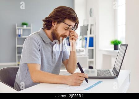 Der müde Mann ist überarbeitet und hat Schmerzen in den Augen, wenn er im Büro am Laptop arbeitet. Stockfoto
