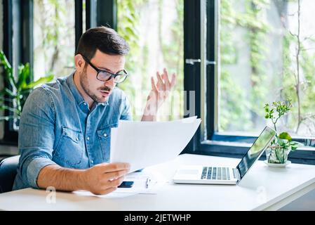 Verwirrt und besorgt Geschäftsmann Analyse von Dokumenten. Seriöse männliche Unternehmer arbeitet auf Laptop am Schreibtisch. Er sitzt im Heimbüro. Stockfoto