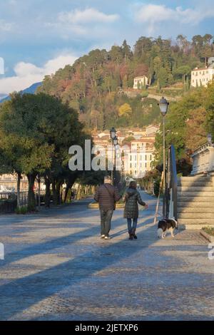 Bellagio, Italien, November 2021. Ein Mann und eine Frau gehen mit ihrem Hund entlang der Promenade von Bellagio am Ufer des Comer Sees. Spaziergänge entlang des Sees. Journe Stockfoto