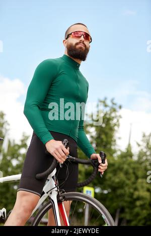 Nahaufnahme eines glücklichen, attraktiven jungen Europäers mit Bart, Brille und grüner Sportkleidung, der beim abendlichen Ausritt im Park wegschaut Stockfoto