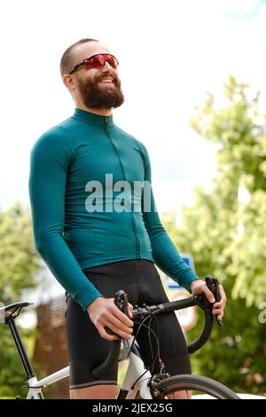 Nahaufnahme eines glücklichen, attraktiven jungen Europäers mit Bart, Brille und grüner Sportkleidung, der beim abendlichen Ausritt im Park wegschaut Stockfoto